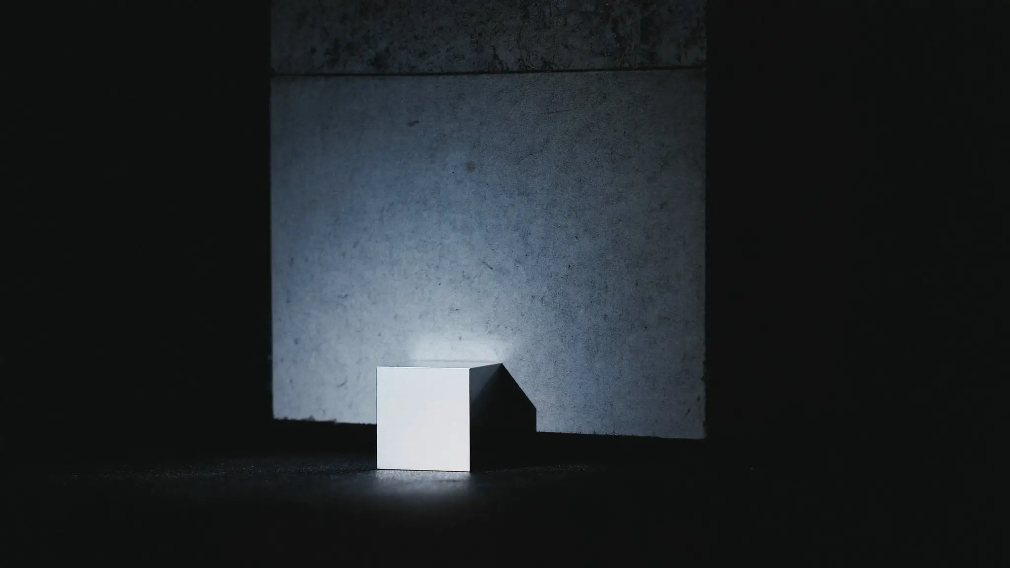 Um pequeno cubo de metal polido iluminado em primeiro plano, ao lado de um bloco de concreto muito maior que se perde na sombra.