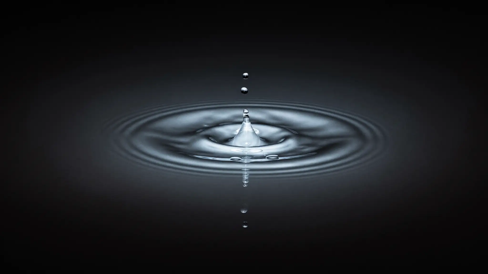 Uma gota atinge uma vasta superfície líquida escura, formando anéis concêntricos que se expandem ao longe.
